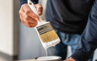 Professional painter's hand holding a paintbrush loaded with fresh white paint, dripping into a can.