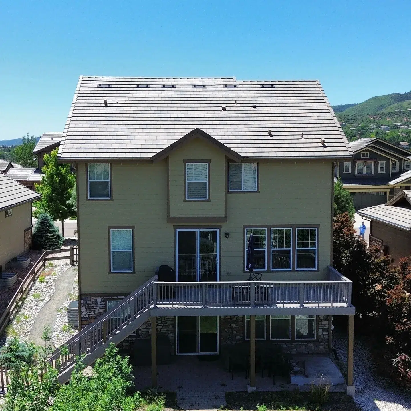 Sage Green Two Story Residential Home Sage green two-story residential home with dark metal roof, black-trimmed windows, stone foundation, and gray deck railing
